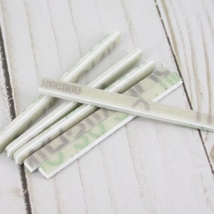 May include: A collection of rectangular, off-white adhesive strips with a camouflage pattern and the word "RECNOW" printed on them. The strips are arranged on a light wood-grain surface.