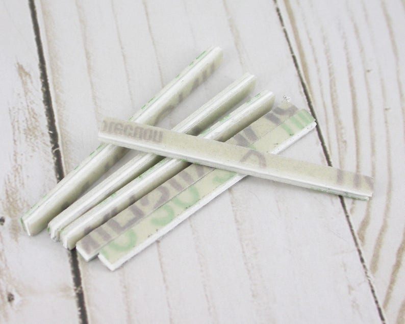 May include: A collection of rectangular, off-white adhesive strips with a camouflage pattern and the word "RECNOW" printed on them. The strips are arranged on a light wood-grain surface.