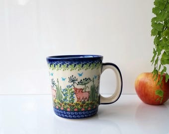 May include: A ceramic mug with a whimsical design featuring pink pigs in a field of flowers and greenery. The mug has a blue rim and handle, and the design wraps around the body of the mug.
