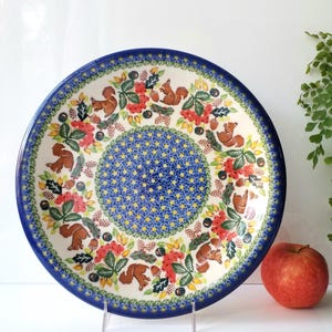 May include: A round ceramic plate with a blue rim and a detailed pattern. The plate features a central blue design surrounded by a floral and squirrel motif in red, green, and brown. An apple rests nearby.