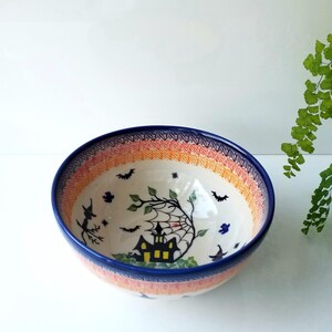 May include: A ceramic bowl featuring a hand-painted Halloween scene with a black haunted house, a spider web, bats, and a witch on a broomstick. The interior rim is decorated with orange and blue patterns, and the exterior has a blue rim.