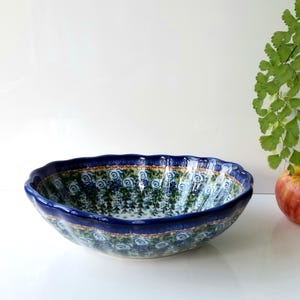 May include: A ceramic bowl with a scalloped edge and a dark blue rim. The bowl features a floral pattern in shades of blue and green. An apple and greenery are in the background.