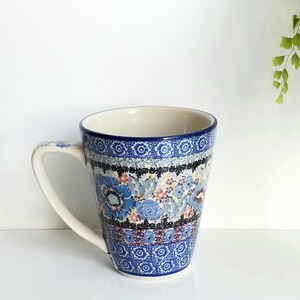 May include: A ceramic mug featuring a cream-colored interior and a blue, white, and black floral pattern. The exterior displays intricate bands of geometric shapes and detailed blue flowers with small pink accents, complemented by a curved handle.