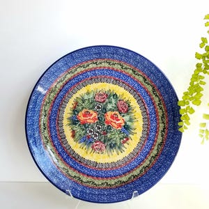 May include: A round ceramic plate featuring a central yellow medallion decorated with a bouquet of red and orange roses and small blue flowers. The border consists of concentric rings in shades of blue, green, and black with intricate patterns.