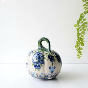 May include: A ceramic pumpkin-shaped decorative object with a cream-colored base, featuring hand-painted clusters of dark blue berries, green leaves, and small light blue flowers. It has a prominent, curved green stem handle on top.