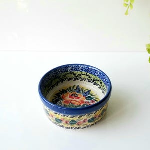May include: A round ceramic bowl featuring a cream-colored base with a hand-painted floral pattern in shades of orange, blue, and green. The rim is decorated with a dark blue border and a delicate black vine motif, while the exterior displays a repeating floral garland design.