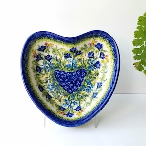 May include: A heart-shaped ceramic dish with a blue and green floral design. The dish features a central heart shape with a blue and white floral pattern surrounded by a border of green leaves and yellow flowers.