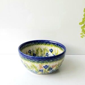 May include: A ceramic bowl featuring a hand-painted pattern of blue flowers, green foliage, and small orange insects. The bowl has a deep blue rim and a cream-colored base, with the floral design wrapping around both the exterior and interior surfaces.