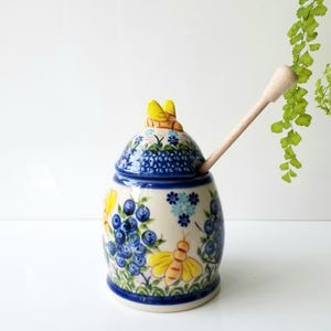 May include: A ceramic honey pot with a lid featuring a yellow bee handle. The pot is decorated with hand-painted blue blueberries, green leaves, and yellow bees on a cream background. A light-colored wooden honey dipper rests inside the pot.