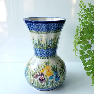 May include: A ceramic vase with a bulbous base and flared top. The vase features a floral design with irises in shades of yellow, purple, and blue, set against a white background. Blue patterned bands encircle the vase.