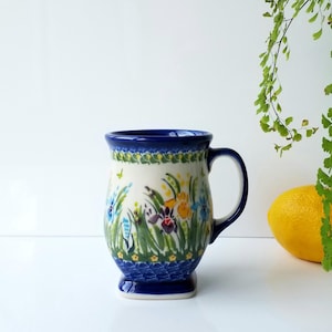 May include: A ceramic mug with a blue handle and base, featuring a floral design with blue, purple, yellow, and green accents. The mug has a white background and a dark blue rim.