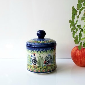 May include: A ceramic jar with a lid featuring a hand-painted pattern of black and white spotted chickens, green foliage, and blue accents. The jar has a cylindrical body and a rounded knob on the lid, finished with a glossy glaze.