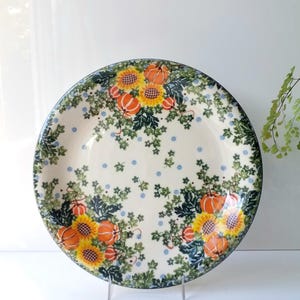 May include: A round ceramic plate with a cream-colored background. The plate features a floral design with sunflowers, pumpkins, and green leaves. The rim is dark blue, and the plate is supported by a clear stand.