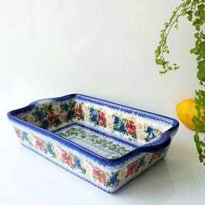 May include: Rectangular ceramic baking dish with a blue rim and handles. The dish is decorated with a floral pattern featuring red, blue, and white flowers on a cream background. A decorative kitchenware item.