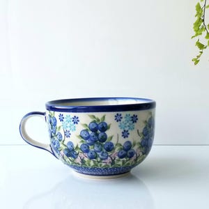 May include: A ceramic mug with a blue floral design featuring blueberries and flowers. The mug has a white base, a blue rim, and a curved handle. The design is hand-painted with shades of blue and green.