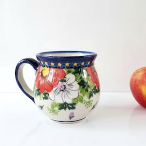 May include: A ceramic mug with a floral design featuring red and white flowers, green leaves, and a blue rim and handle. A red apple sits to the right of the mug. The mug is on a white surface.