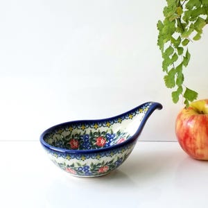 May include: A ceramic spoon rest with a blue rim and handle, decorated with red and blue floral patterns. The spoon rest is on a white surface, with a red and yellow apple and green foliage in the background.