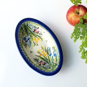 May include: An oval ceramic bowl with a deep blue rim and a cream-colored interior featuring a hand-painted floral pattern of yellow, blue, and dark purple irises with green stems and leaves, finished with a decorative blue sponge-stamped border.