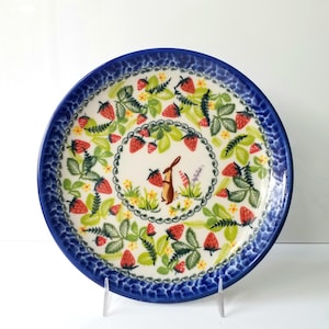 May include: A round ceramic plate with a blue rim and a strawberry design. The plate features a central illustration of a brown rabbit holding a strawberry, surrounded by red strawberries and green leaves. The plate is supported by a clear stand.
