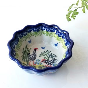 May include: A small, white ceramic bowl with a blue rim and a scalloped edge. The bowl is decorated with a colorful floral pattern and two chickens.