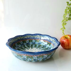 May include: A ceramic serving bowl with a scalloped edge, featuring a blue and green floral pattern. The bowl is next to a red and yellow apple. The bowl's rim is a dark blue.