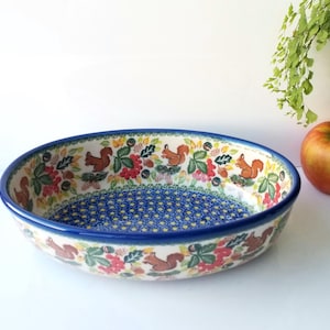 May include: Oval ceramic serving bowl with a blue rim and a detailed pattern. The bowl features a blue dotted interior and a border with squirrels, leaves, and flowers in red, green, and brown. An apple and greenery are in the background.