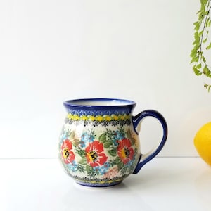May include: A ceramic mug with a blue handle and rim, adorned with a floral pattern featuring red poppies, blue and green leaves, and yellow accents. The mug is set against a white background with a lemon and greenery.