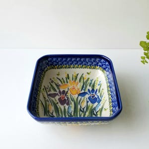 May include: A square ceramic bowl with a cream-colored interior featuring a hand-painted floral design of blue, purple, and yellow irises with green stems. The rim is painted in a solid dark blue, and the exterior edge has a blue patterned border.