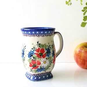May include: A ceramic mug with a white base, adorned with red poppies, blue flowers, and green leaves. The rim and base feature a blue decorative pattern. A red and yellow apple sits beside the mug.