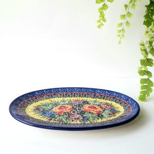 May include: An oval ceramic platter featuring a vibrant floral pattern with orange and pink roses, green leaves, and blue accents on a yellow background. The rim is decorated with a dark blue and black intricate geometric border design.