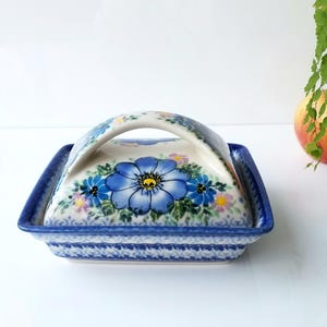May include: A square ceramic butter dish with a handle, featuring a blue floral design on a white background. The dish has a blue border and a matching lid with a curved handle. The floral pattern includes large blue flowers and smaller colorful accents.