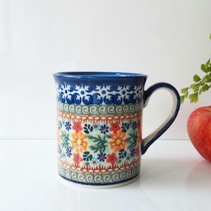 May include: A ceramic mug with a blue handle and rim, adorned with a floral pattern in red, orange, and blue. The mug is approximately 4 inches tall and sits on a white surface. An apple and greenery are in the background.