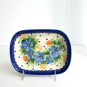 May include: A rectangular ceramic dish with rounded corners, featuring a cream-colored base decorated with blue and orange floral patterns, green foliage, and small blue hearts, finished with a dark blue circular border pattern along the rim.