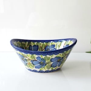 May include: A ceramic serving bowl with a blue floral design. The bowl has a curved shape with a dark blue rim and a scalloped edge. The interior features blue flowers and green leaves on a white background.