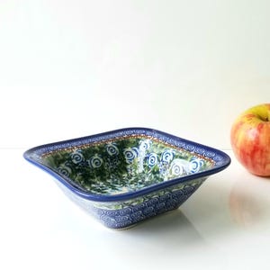 May include: Square ceramic bowl with a blue rim and floral pattern. The bowl features a green and blue design with white swirl details. A red and yellow apple sits next to the bowl.