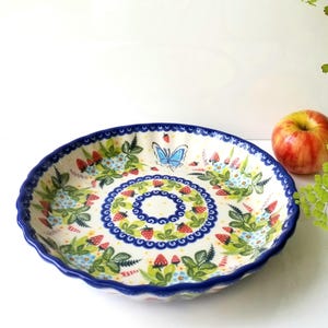 May include: A round ceramic serving dish with a scalloped edge and a blue rim. The dish features a colorful floral and butterfly design with red strawberries, green leaves, and blue accents. An apple rests beside the dish.