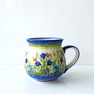 May include: A ceramic mug with a blue rim and handle, featuring a floral design with blue flowers and yellow insects. The mug has a white interior and a rounded shape, perfect for enjoying hot beverages.