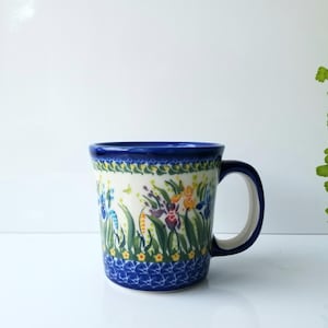 May include: A ceramic mug featuring a cream-colored background with a hand-painted floral pattern of blue, yellow, and purple flowers amidst green foliage. The rim and base are decorated with a dark blue border with a repeating white pattern.