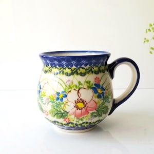 May include: A ceramic mug with a rounded shape and a large handle. The mug features a floral design with pink, blue, and green flowers on a cream background. The rim and handle are dark blue, and the mug is approximately 4 inches tall.