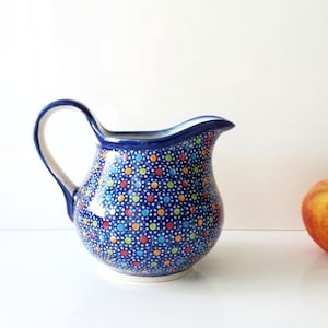 May include: A blue ceramic pitcher with a white handle and a spout. The pitcher is decorated with a pattern of colorful dots. An apple is next to the pitcher.