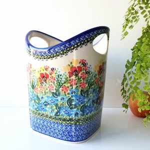 May include: A ceramic utensil holder with a floral design. The holder is white with a blue rim and base, and features a colorful pattern of red, blue, and yellow flowers. It has two handles.