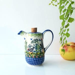 May include: A ceramic pitcher with a cork stopper. The pitcher features a hand-painted design of birds, flowers, and foliage in blue, green, and red on a white background. It has a curved handle and a spout. An apple and greenery are in the background.
