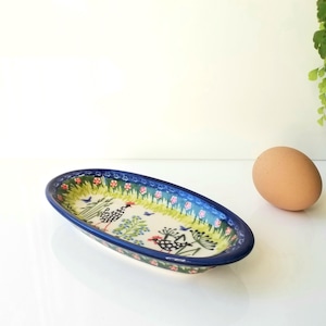 May include: An oval ceramic dish with a dark blue rim, featuring a hand-painted pattern of black and white chickens, green foliage, and small red flowers on a cream background, positioned next to a single brown egg on a white surface.