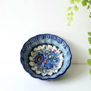 May include: A round ceramic bowl with a scalloped edge, featuring a detailed floral pattern in shades of blue, white, and black. The interior displays a central cluster of blue flowers surrounded by intricate decorative borders and foliage.