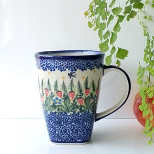 May include: Tall ceramic mug with a blue and white floral design. The mug features a handle and a pattern of strawberries, ferns, and a bird. The mug is approximately 6 inches tall.