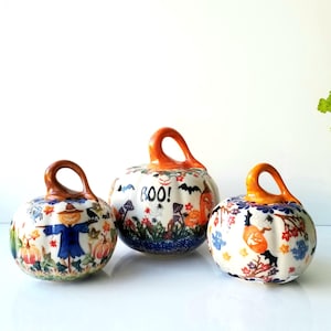 May include: Three ceramic pumpkins with orange stems and white bases featuring colorful hand-painted motifs. The center pumpkin displays the text "BOO!" alongside bats, mushrooms, and foliage. The others feature a scarecrow and autumn leaves.
