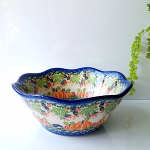 May include: A ceramic bowl with a scalloped rim featuring a colorful pattern of orange pumpkins, green leaves, and small dark berries against a cream background, finished with a deep blue painted border along the top edge and base.