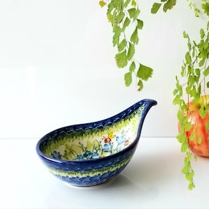 May include: A blue and green ceramic spoon rest with a floral and bee design. The spoon rest has a curved handle and a wide, shallow bowl. The rim is a dark blue, and the interior features a yellow and green floral pattern.