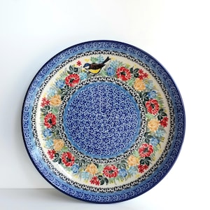 May include: A round ceramic plate featuring a central blue and white speckled pattern surrounded by a decorative border of red poppies, yellow roses, and blue flowers, with a small bird perched among the floral design on a white background.
