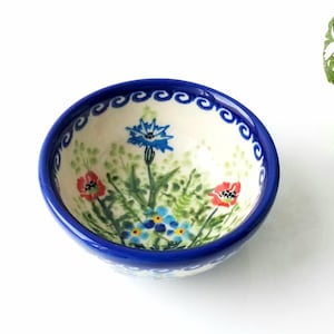 May include: A small, white ceramic bowl with a blue rim and a floral design. The bowl features blue, red, and green flowers on a white background.